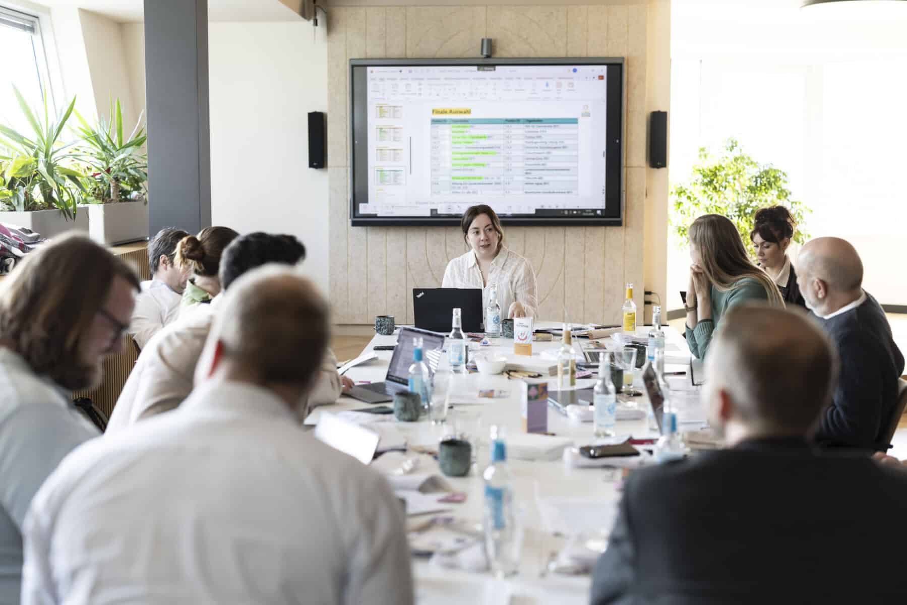 10 Personen sitzen um einen Tisch herum und diskutieren. Im Hintergrund ist ein Fernseher mit Tabellen zu sehen.