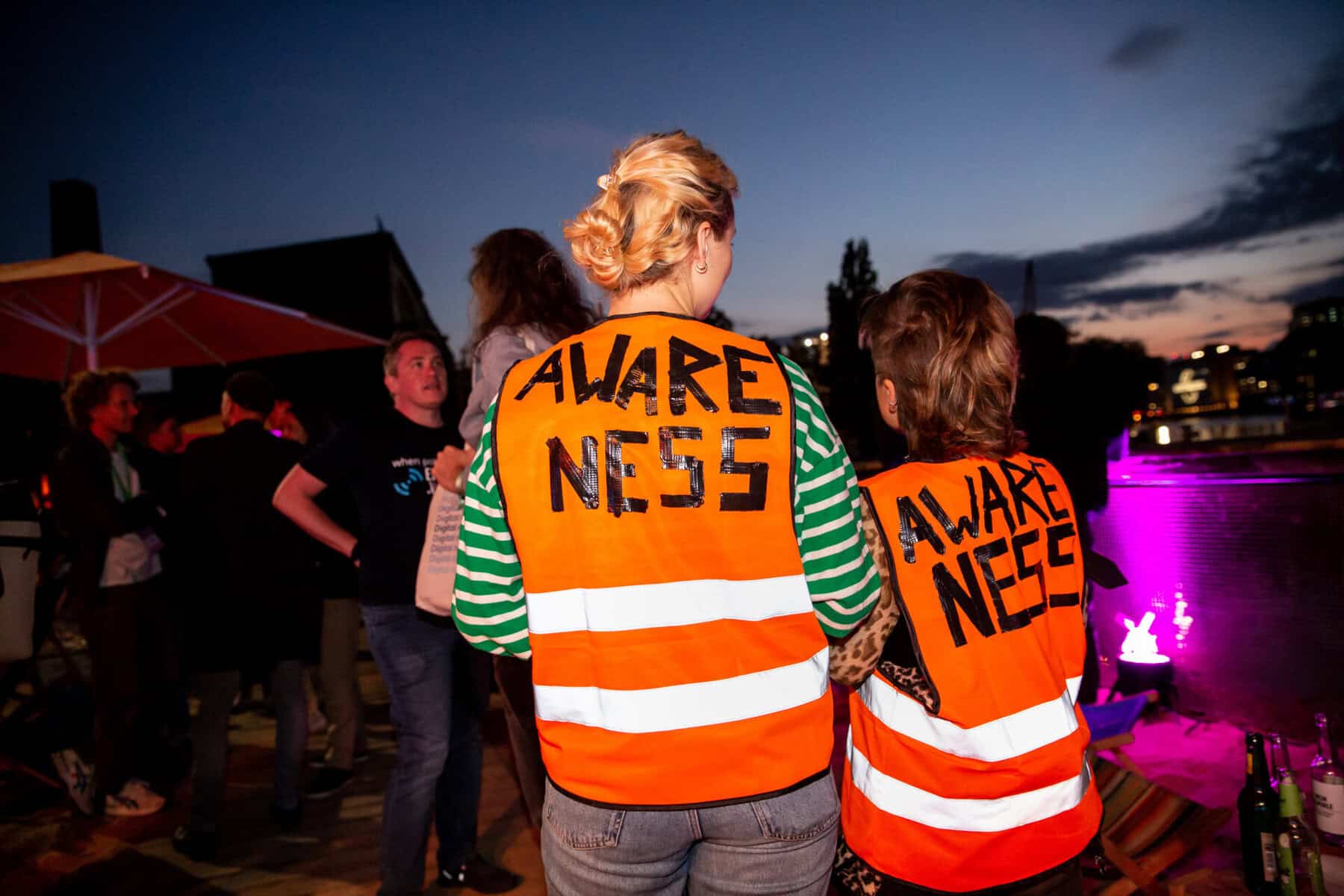 Menschenmende mit zwei Personen im Vordergrund die orangene Westen mit der Aufschrift "Awareness" tragen