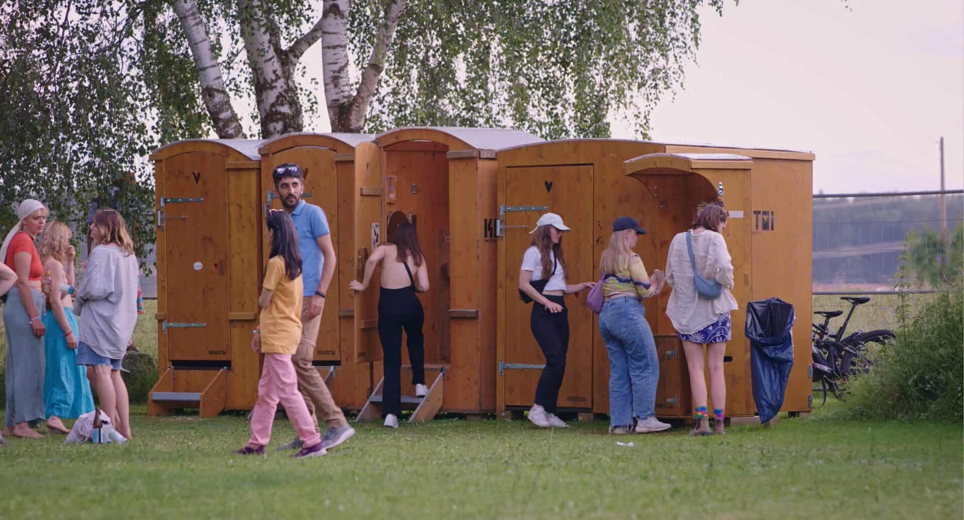 Auf einer sommerlichen Wiese steht eine Reihe Komposttoiletten und ein Handwaschbecken. Es gehen Personen rein und raus, warten oder waschen sich die Hände.