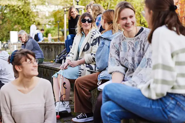 Eine Gruppe Menschen sitzen zusammen und unterhalten sich, darunter auch eine Frau mit Blindenstock und Sonnenbrille.