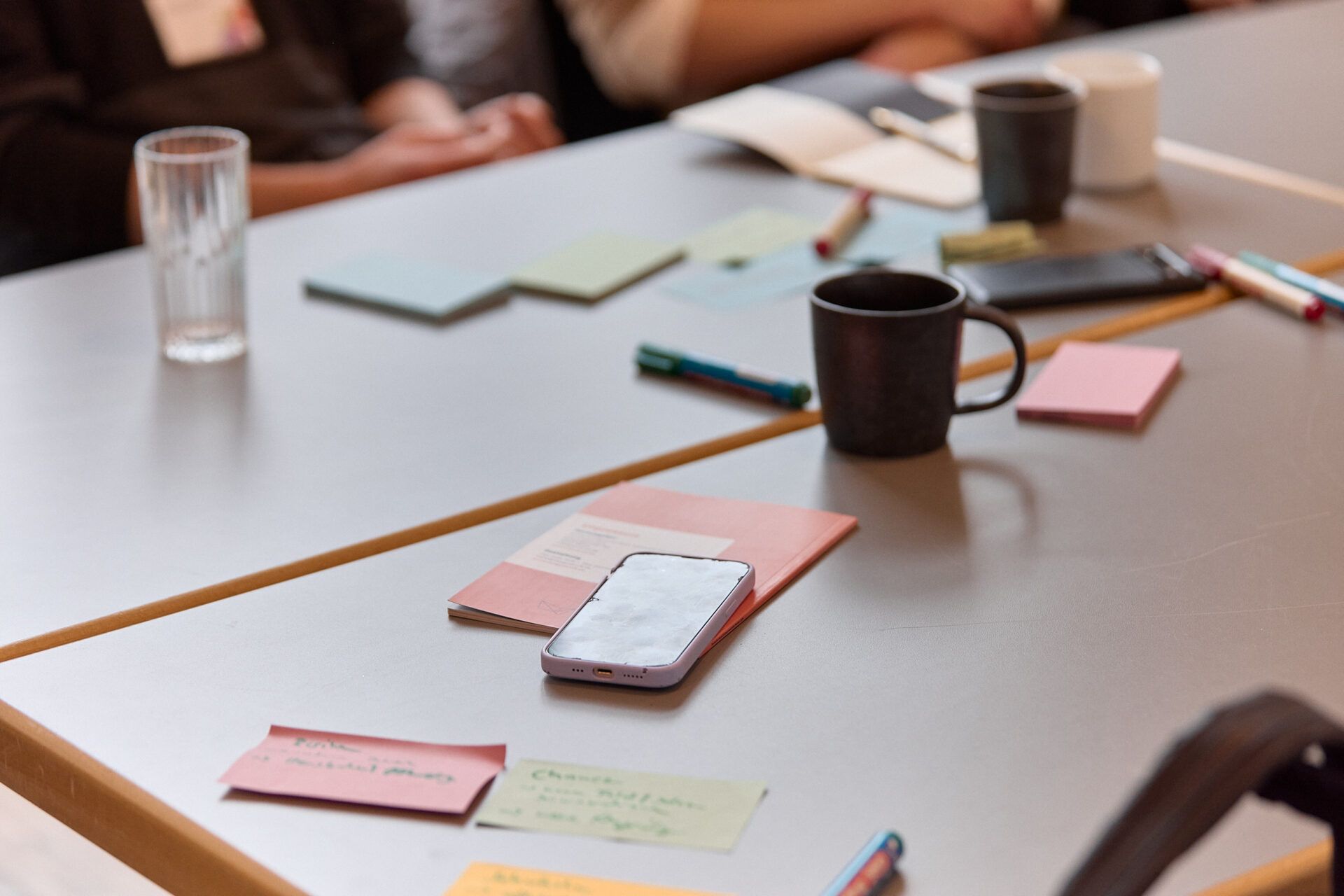 Bild von einem Tisch mit Postits, Stiften und Tassen. Menschen hinten nur unscharf zu erkennen.