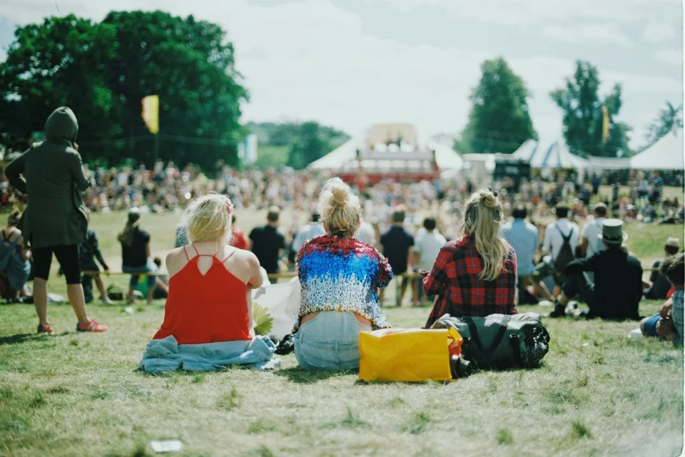 drei Frauen sind mit dem Rücken zu sehen und tragen sommerliche, bunte Outfits. Vor ihnen ein Festivalgelände