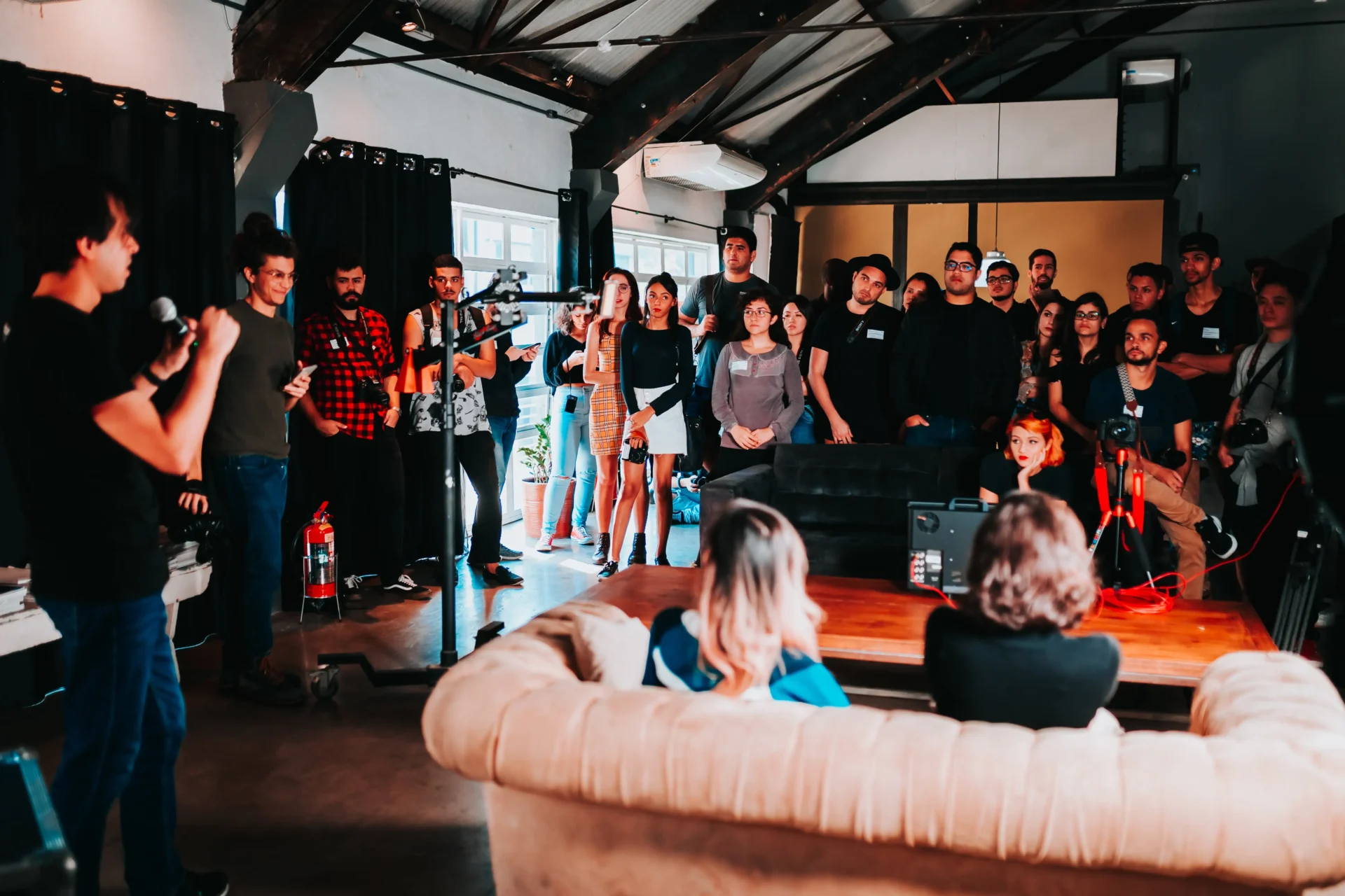 Links im Bild steht ein Mann mit einem Mikrofon und erzähl einer Runde von ca. 20 Personen etwas. Ihn Raum ist auch ein Sofa von hinten zu sehen und Kameras, die auf den Sprecher gerichtet sind.