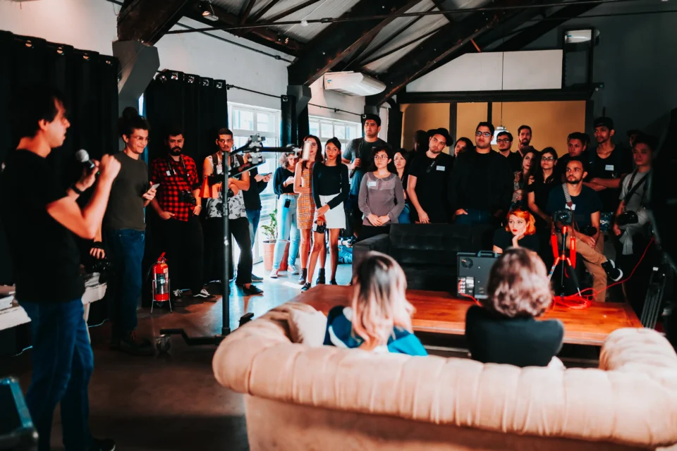 Links im Bild steht ein Mann mit einem Mikrofon und erzähl einer Runde von ca. 20 Personen etwas. Ihn Raum ist auch ein Sofa von hinten zu sehen und Kameras, die auf den Sprecher gerichtet sind.